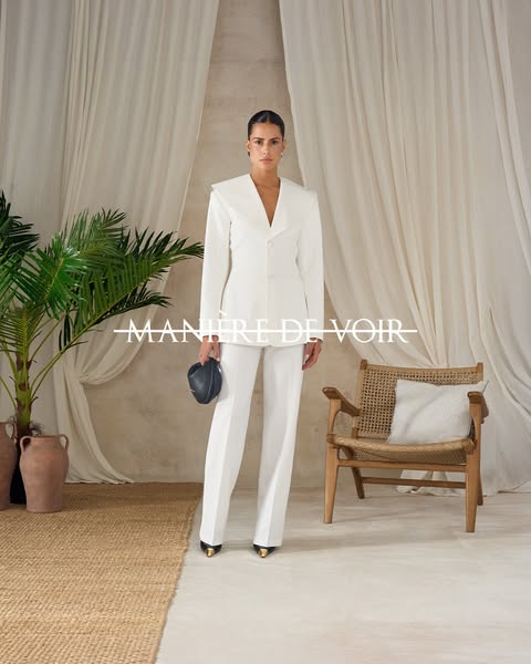 This ad features a woman wearing a white suit, standing in front of a tan-colored wall and light beige drapery. The bran