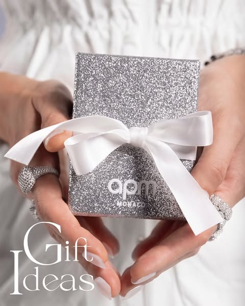 This image ad showcases a gift idea from APM Monaco, featuring a silver glittery box tied with a white ribbon, held by a