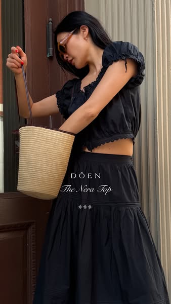 A woman in a black outfit poses outside near a door. The ad is for the Nera Top from Doen.