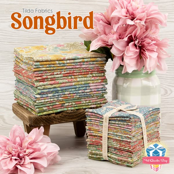This ad features a stack of Tilda Fabrics Songbird fat quarters set on a wooden table. Pink flowers are in the backgroun
