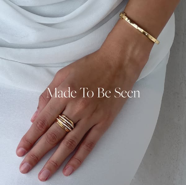 This image ad showcases gold jewelry, specifically a ring and bracelet, worn by a person with tan skin against a light-c