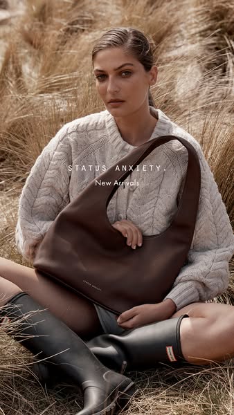 This Status Anxiety ad features a model seated in a field of tall grass holding and showcasing a brown leather handbag. 