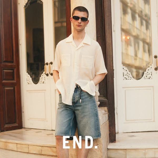 An image ad featuring a man in a light-colored shirt and jean shorts in front of a building with white doors. The brand 