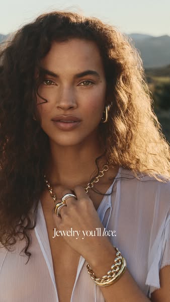 A close-up shot of a model wearing several gold jewelry pieces. The jewelry is in focus and the background is a blur of 