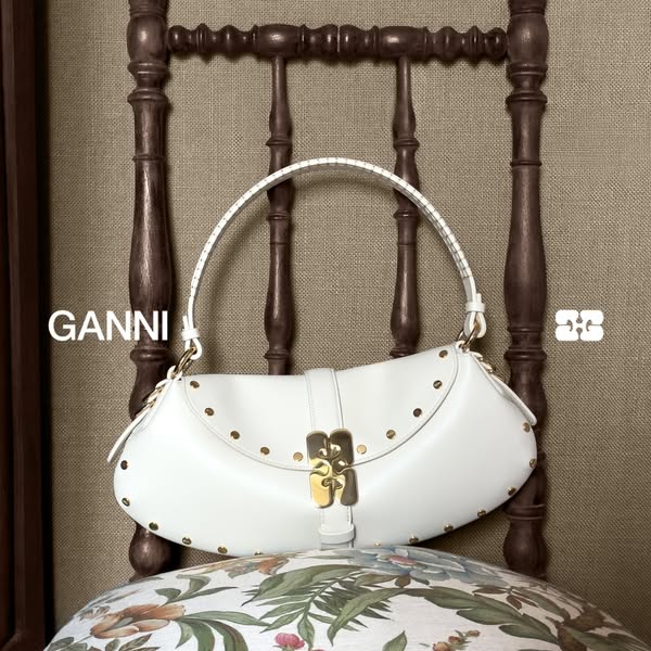 This image ad features a white GANNI handbag with gold hardware, styled on a vintage chair to highlight the brand's 