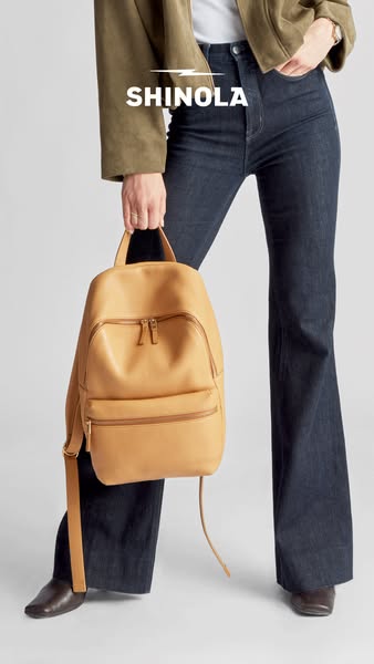 An image ad for a Shinola backpack. The product is shown being held by someone whose torso and legs are visible. The mod