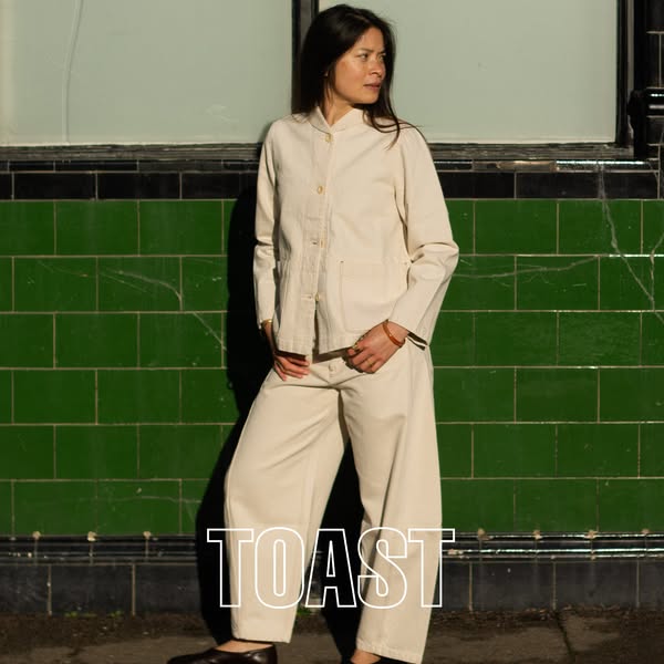 A woman poses wearing matching separate pieces from Toast. The pieces are a matching jacket and pants in an off-white co