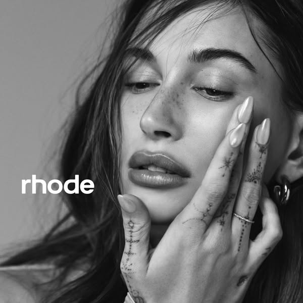 A black and white image of Hailey Bieber with her hand on her face. The brand name appears on the left.