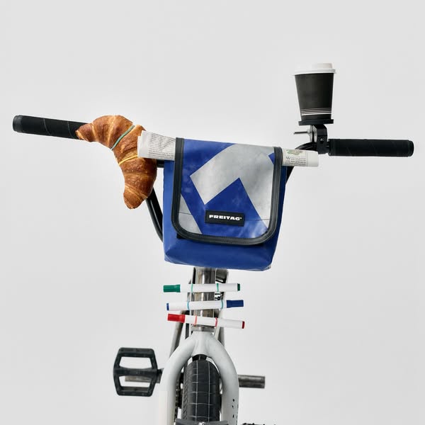 This ad is displaying the FREITAG brand messenger bag attached to a bicycle with a newspaper, croissant, and coffee atta