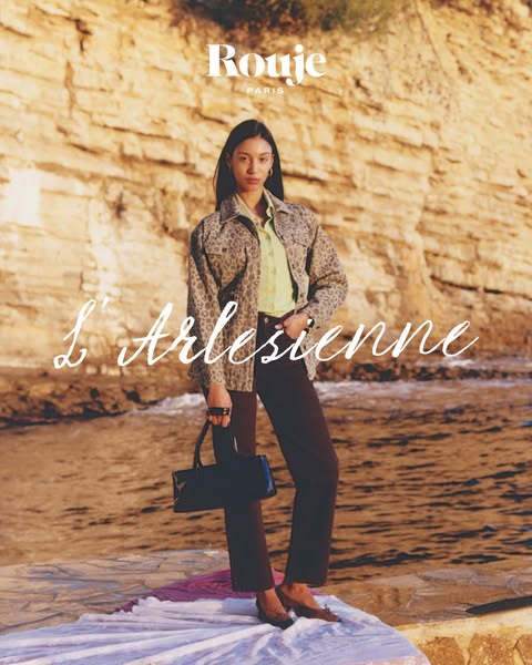 This ad features a model wearing a Rouje Paris brand jacket, blouse, pants, shoes, and handbag, standing on a white clot