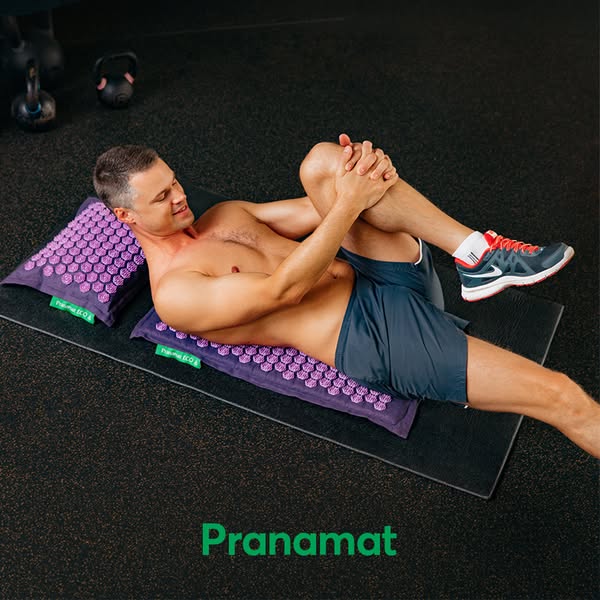 A man is lying on a spike mat while stretching his leg in a gym setting.