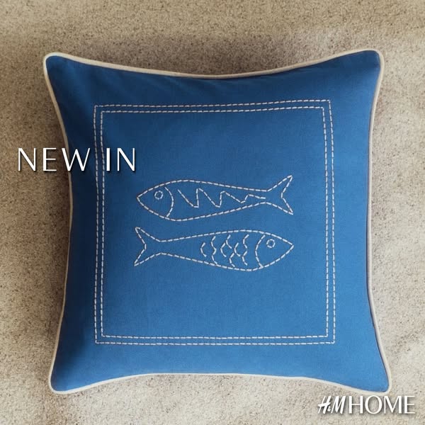 This is a static image promoting a new decorative pillow from H&M Home. The pillow is blue with a white embroidered 