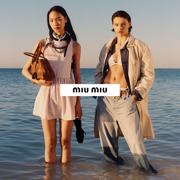 An image ad showcases two models standing in shallow water in a coastal setting. The models wear clothing and accessorie