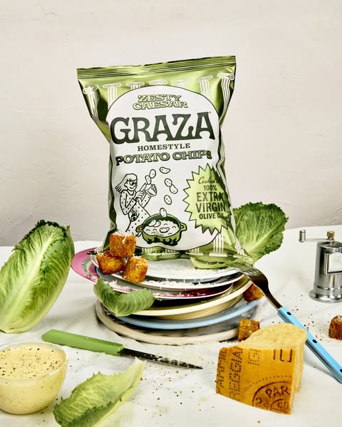 This ad features a bag of Graza Zesty Caesar Homestyle Potato Chips arranged amidst the ingredients and elements of a ca