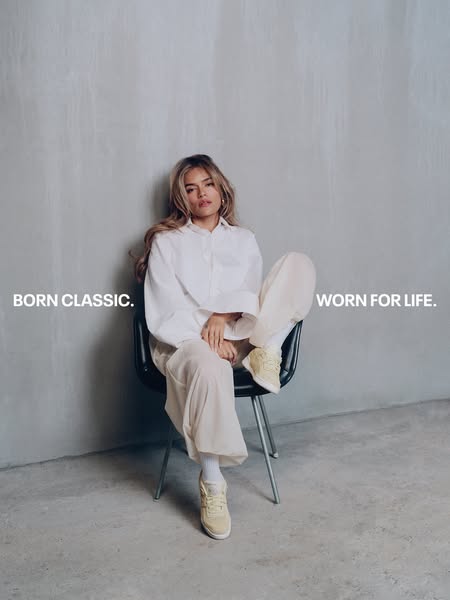 This image ad shows a woman wearing a white top and white pants sitting in a black chair wearing sneakers to advertise s