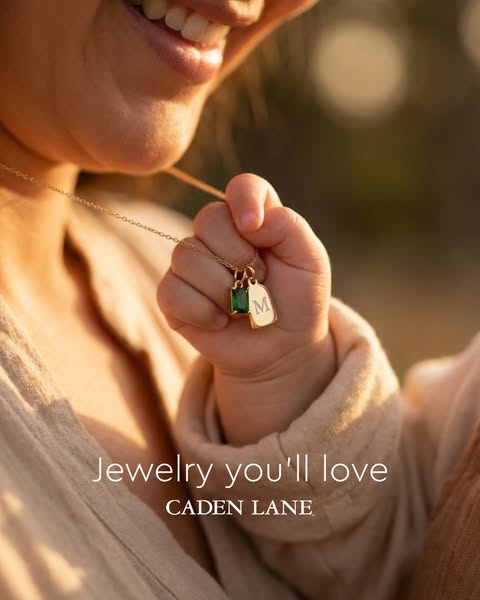 This image ad for Caden Lane jewelry features a close-up of a baby's hand holding a gold necklace. The necklace has 
