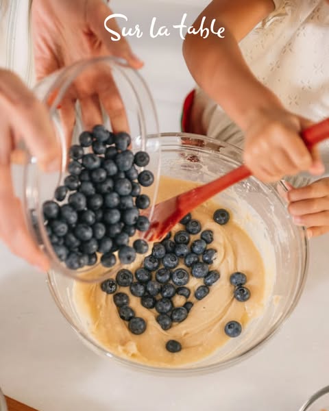 An overhead shot of a child and an adult making a dessert with blueberries in a clear bowl.