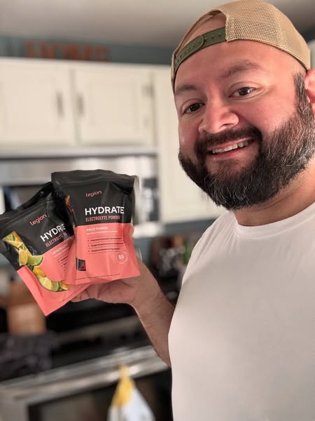 A man in a kitchen setting holds up two packages of Legion Hydrate electrolyte powder and smiles at the camera.