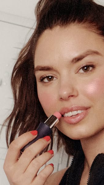 A woman applies a rose colored lip balm to her lips. She is looking directly at the camera and showing off the color and