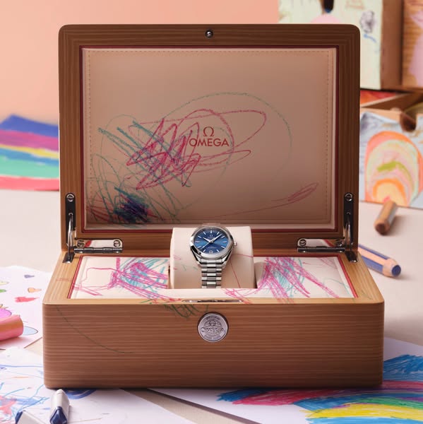 This image ad features an Omega watch displayed in an opened wooden box covered with colorful crayon scribbles. The pres
