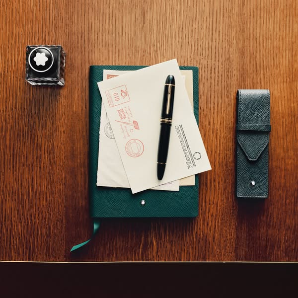 This ad showcases a Montblanc pen and ink set on a wooden desk next to a closed green notebook.