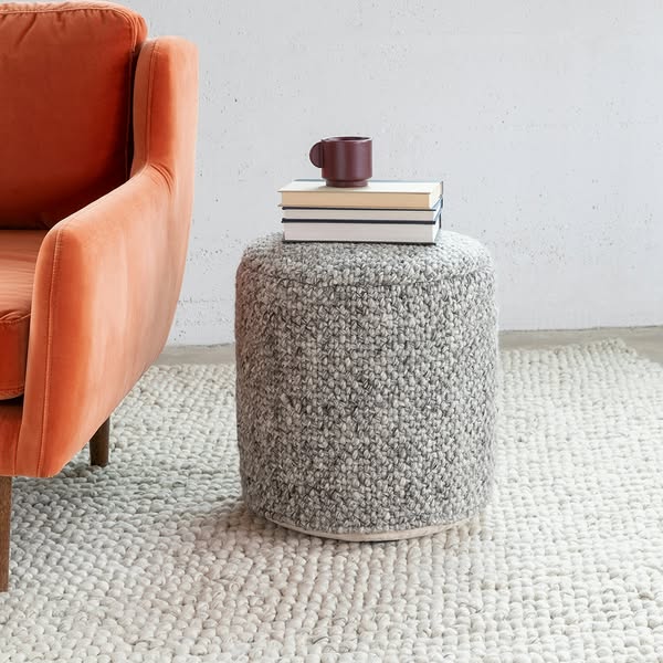 This ad features a knitted grey ottoman with a stack of books and a mug on top, next to an orange chair on a white rug.
