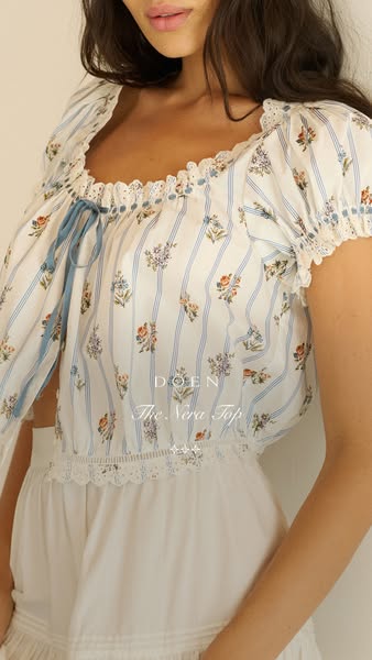 A close-up shot of a white and blue floral blouse with lace trim on a fair-skinned model.  The brand and product name ar