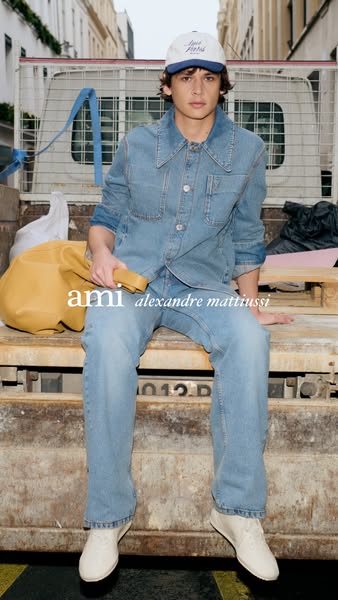 A male model in double denim from Ami Paris sits on the back of a truck, wearing a baseball cap and cream sneakers, hold