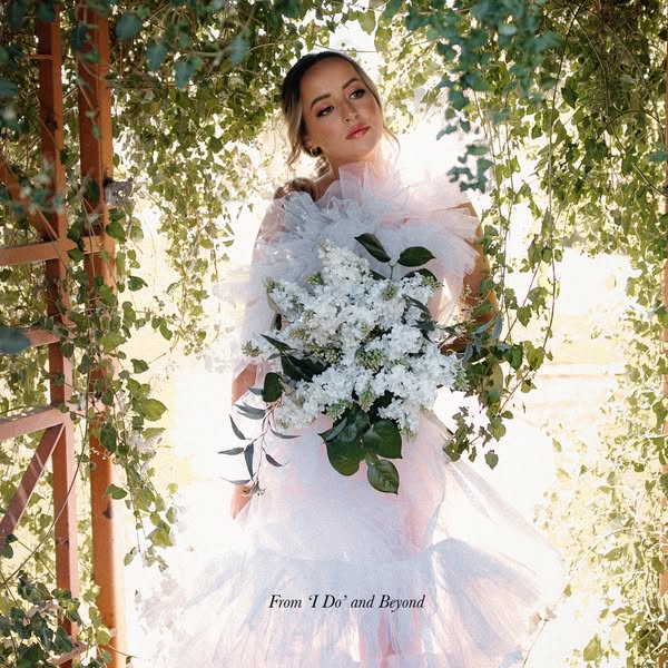 An image featuring a person in a white dress holding a bouquet of flowers in a natural setting, promoting 'I Do'