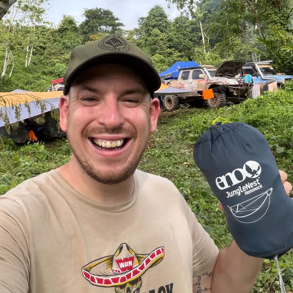 A smiling man with a hat and t-shirt stands outdoors in front of off-road vehicles, holding the ENO JungleNest Hammock. 