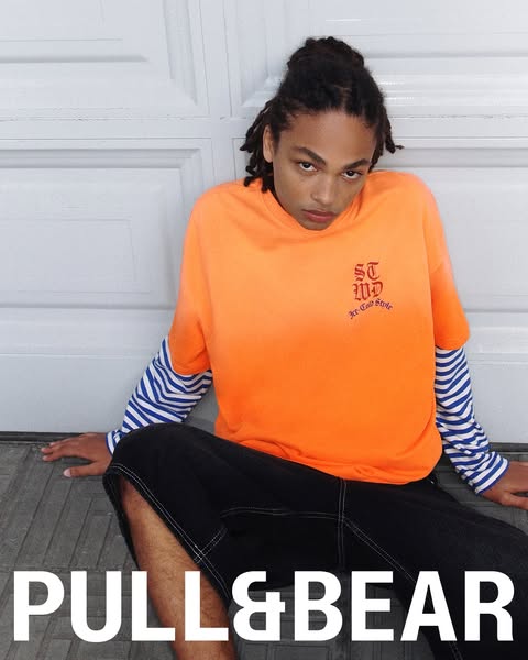 This is an image ad by Pull&Bear, featuring a model sitting against a white door wearing an orange t-shirt and black