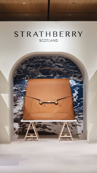 This ad features a Strathberry handbag prominently displayed in a bright, minimalist setting. The focus is on showcasing