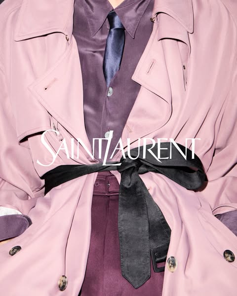 A close-up of a lavender trench coat tied with a black satin belt. The Saint Laurent logo is displayed across the midsec