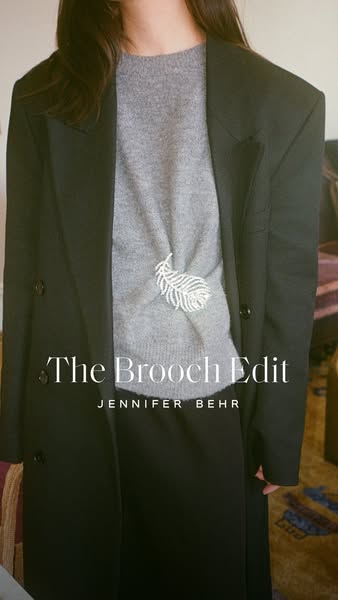 This image promotes the 'Brooch Edit' collection by Jennifer Behr, showcasing a single brooch on a sweater and c