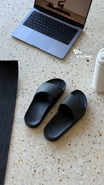 This ad shows a pair of black slides sitting on a speckled floor, with a laptop and yoga mat in the background to demons
