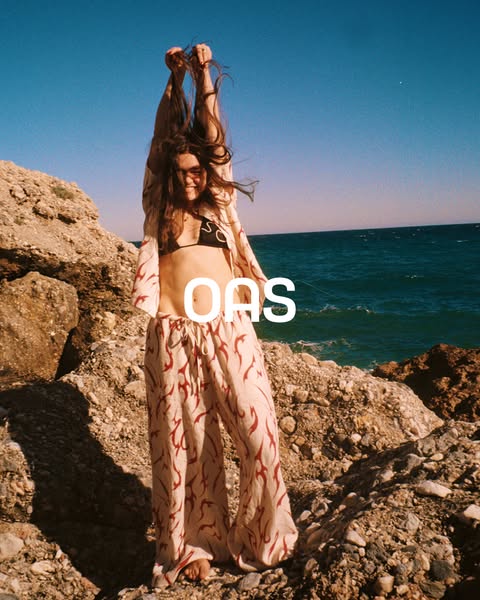 A woman stands on a rocky beach wearing matching shirt and pants. The brand logo "OAS" is overlaid on the imag