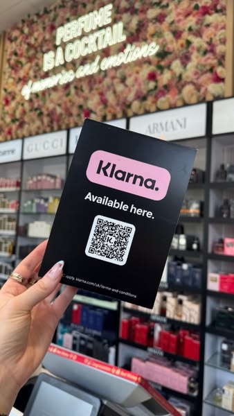 This image shows a black sign with the Klarna logo and a QR code on it, held in front of a shelf of perfume bottles in a