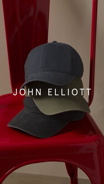 This image ad showcases three John Elliott baseball caps stacked on a red chair. The brand name is displayed prominently