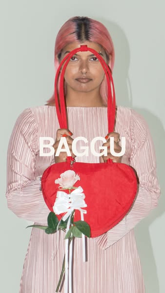 An image features a person holding up a heart-shaped red BAGGU bag adorned with a white ribbon and a rose.