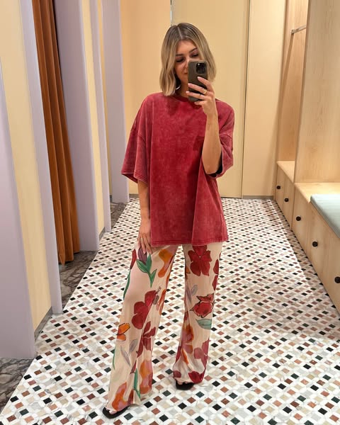 A woman poses in a fitting room for a selfie in a red t-shirt and floral pants.