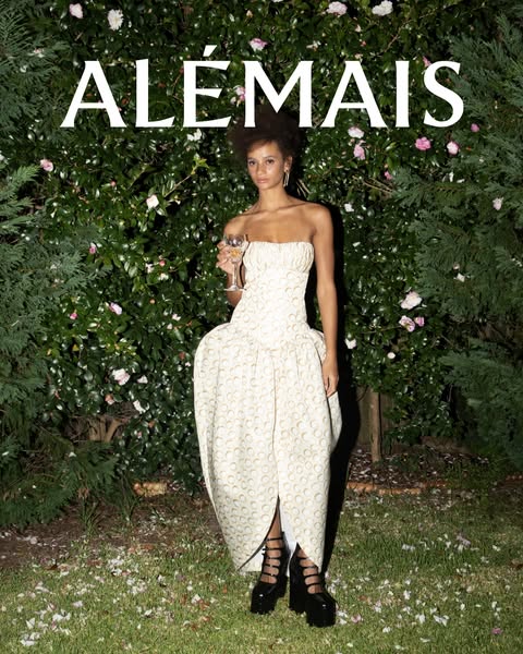 This image ad for ALÉMAIS features a model wearing a cream-colored dress with gold detailing, standing in a garden setti