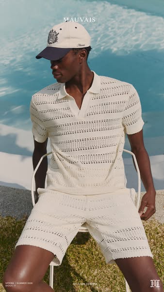 A medium shot of a male model sitting outside near a pool, wearing a matching knitted polo shirt and shorts set. A baseb