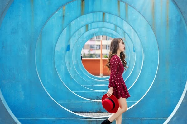 This lifestyle image features a young woman in a fashionable red dress walking through a blue tunnel, holding a red hat.