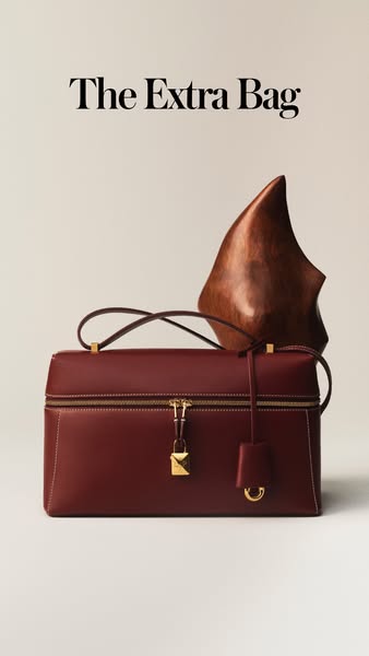 A minimalist product shot featuring a red leather bag. The bag is displayed with a wooden shoe form placed behind it on 
