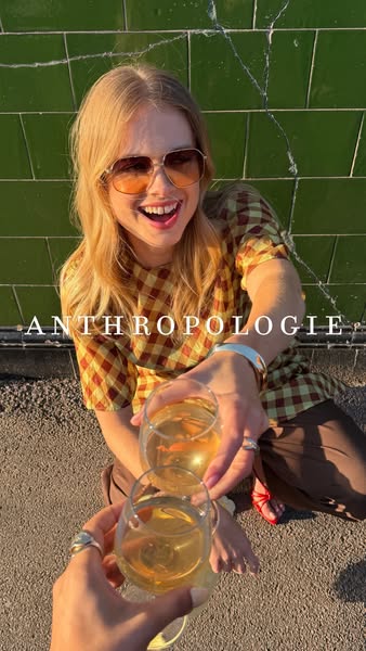 This image shows a young woman toasting wine glasses with another person off screen while dressed in Anthropologie cloth
