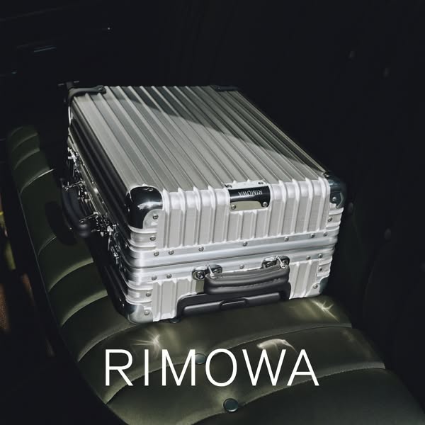 This image ad shows a silver RIMOWA suitcase on a cushioned seat in a dark environment, highlighting the brand name.