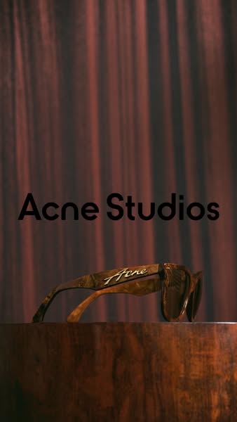 This ad showcases Acne Studios sunglasses on a wood surface with a brown background curtain.
