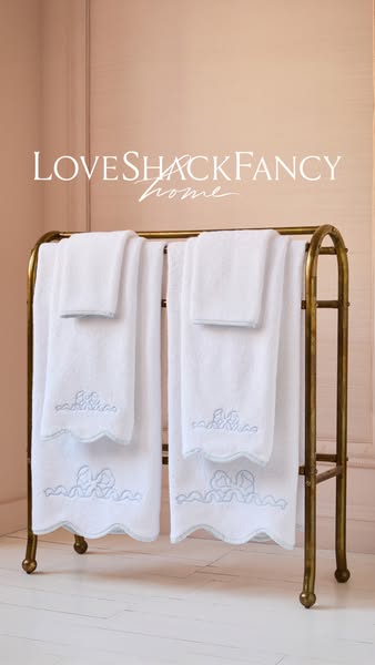 This image ad showcases a set of LoveShackFancy Home towels on a brass towel rack against a neutral wall. The towels are