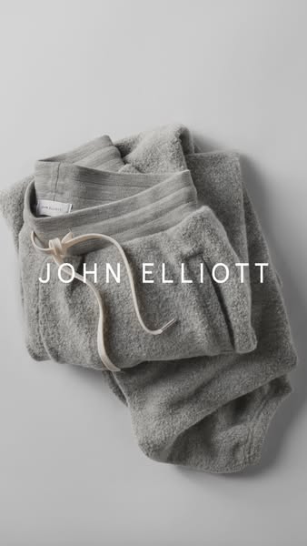 This ad showcases a pair of grey John Elliott sweatpants. The image is a close-up shot of the pants folded on a light gr