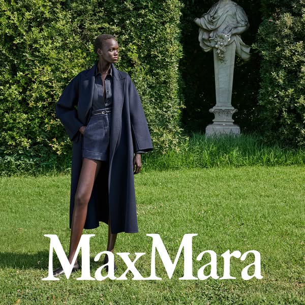 This ad features a woman in a dark blue outfit and coat standing on a lawn in front of a hedge and statue. The brand nam
